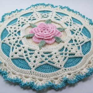 Op de afbeelding: Een wit gehaakt doily met een blauw midden en een roze gehaakte bloem in het midden.