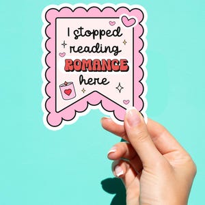 Puede incluir: Una pegatina rosa con borde festoneado y el texto "I stopped reading ROMANCE here". La pegatina presenta un diseño de banner con corazones, estrellas y una ilustración de vela. La pegatina está sujeta por una mano sobre un fondo turquesa.