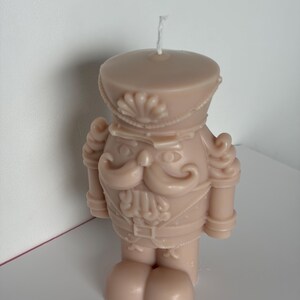 Può includere: Una candela rosa chiaro a forma di soldato schiaccianoci. La candela presenta un design dettagliato, tra cui cappello, baffi e uniforme. La candela è in piedi su una superficie bianca.