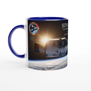 May include: A ceramic mug with a dark blue handle and interior, featuring a graphic of the Apollo-Soyuz spacecraft in orbit above Earth. The design includes the mission patch, the text SOYUZ, and the words First International Space Mission.