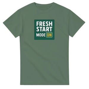 May include: A green t-shirt with a square graphic in the center. The graphic reads "FRESH START MODE ON" in white and yellow lettering on a dark green background. The shirt is made of a soft material.