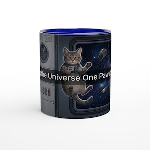 Peut inclure: Mug en céramique avec intérieur bleu et motif enveloppant. Le motif représente un chat tigré gris en combinaison spatiale flottant devant une fenêtre avec un fond étoilé. Le texte sur la tasse se lit "the Universe One Paw."