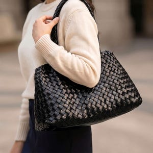 May include: Black woven leather tote bag with a braided handle. The bag has a rectangular shape and a textured surface. The bag is being carried over the shoulder.