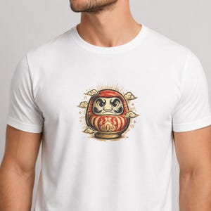 May include: A white short-sleeved t-shirt featuring a centered graphic of a red and gold Daruma doll. The doll has a stern facial expression with thick eyebrows and a mustache, surrounded by stylized golden clouds and radiating light lines.