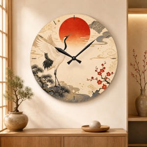 Könnte beinhalten: Eine runde Wanduhr mit cremefarbenem Zifferblatt, das eine große rote Sonne, einen weißen Kranich mit schwarzen Flügelspitzen, stilisierte Kiefern und rote Pflaumenblüten zeigt. Die Uhr hat schwarze Zeiger und einfache schwarze Strichmarkierungen.