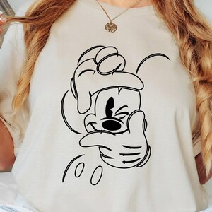 May include: A sand-colored t-shirt featuring a black outline of Mickey Mouse making a hand gesture. The shirt is worn by a person wearing light-wash jeans. The word "SAND" is printed on the upper right corner.