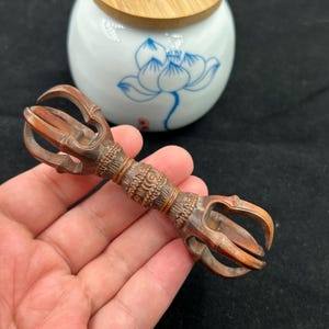 Puede incluir: Un objeto ritual vajra de madera tallada marrón con intrincados patrones geométricos y florales en el mango central, con puntas simétricas en ambos extremos. Al fondo, un tarro de cerámica blanca con un diseño de loto azul y tapa de madera.