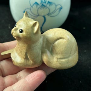 May include: A wooden cat figurine in a resting pose. The cat is carved from light-colored wood, with visible grain and smooth curves. It has small, dark eyes and a pointed ear. The figurine is a decorative item.