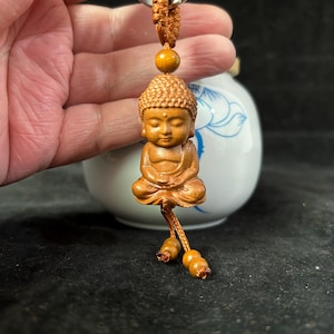 May include: A small, intricately carved wooden Buddha figurine. The seated figure is light brown with detailed facial features and a textured head. It hangs from a knotted cord with a wooden bead and two smaller beads at the end, suggesting a keychain or pendant.