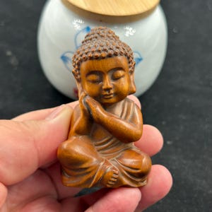 Puede incluir: Pequeña estatua de Buda de madera en posición sentada con las manos juntas en oración. La estatua está tallada en madera de color marrón claro, con rasgos faciales detallados y una expresión serena. El Buda mide unos 5 cm de altura.