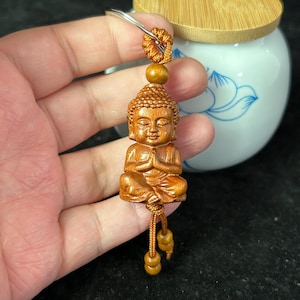 May include: A wooden Buddha keychain with a silver keyring. The Buddha is carved in a seated position with hands in prayer. The keychain is made of light brown wood and has a detailed carving of the Buddha's face and robes.
