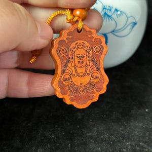 May include: A wooden pendant featuring a carved smiling Buddha. The pendant is a warm orange hue and has a decorative border. It is suspended from a gold-coloured cord.
