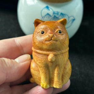 May include: A small, wooden cat figurine with a light brown finish. The cat is carved in a sitting position with a collar and a small, round pendant. The eyes are black, and the cat has a slight smile. A white and blue ceramic jar is in the background.