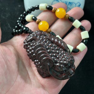 May include: A dark brown carved pendant necklace featuring a detailed mythical creature design. The necklace includes a beaded strand with black, cream, and yellow beads. The pendant is held in a hand.