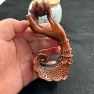Puede incluir: Escultura de madera tallada de una mano sosteniendo una flor de loto. La mano está detallada con pulseras y patrones intrincados. La escultura es de color marrón cálido.