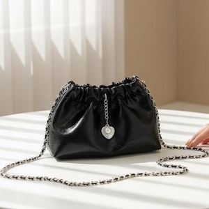 Puede incluir: Bolso de hombro de cuero negro con una correa de cadena plateada. El bolso tiene una parte superior fruncida y un pequeño dije en forma de corazón con un acento de perla. El bolso está sobre una superficie blanca.