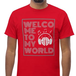 以下が含まれることがあります： グレーの長方形のフレームに「WELCOME TO MY WORLD」という文字が入った赤いTシャツ。デザインには、ヘッドホンと音符をつけた脳が含まれています。モデルはブルージーンズを着用しています。
