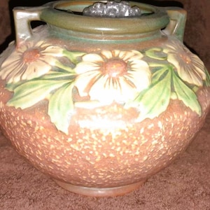 May include: A brown ceramic vase with a textured surface and a green rim. The vase is decorated with embossed white and orange daisies and green leaves. It has two handles and a decorative top.