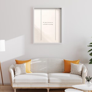 May include: A framed print with the text "the space between us still holds everything" in a minimalist style. The print hangs above a white sofa with orange and gray throw pillows. A round woven rug and small white tables complete the living room scene.
