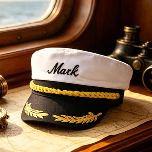 May include: A white and black captain's hat featuring the name Mark embroidered in black script on the front, accented with a gold braided cord and gold leaf embroidery on the brim, resting on a vintage map with nautical navigation tools.