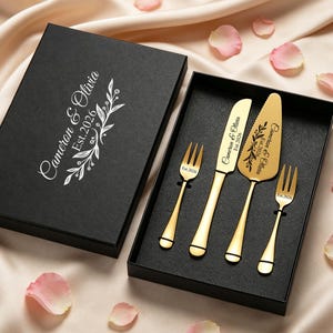 Puede incluir: Un juego de servir pastel de bodas dorado, que incluye dos tenedores, un cuchillo y una espátula, presentado en una caja negra. El juego está grabado con "Cameron & Olivia Est. 2026" y detalles florales. La caja está abierta.