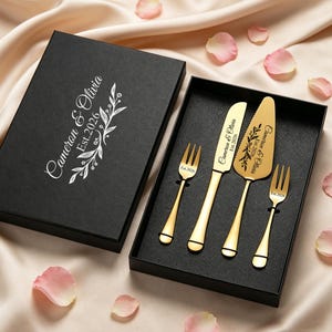 May include: A gold wedding cake serving set, including two forks, a knife, and a server, in a black box. The box and utensils are engraved with the names "Cameron & Olivia" and the date "Est. 2026". The set is presented on a satin surface with scattered rose petals.