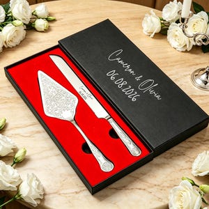 Op de afbeelding: Een zilveren taartmes- en serveerset met sierlijke details, gepresenteerd in een zwarte doos met rode voering. De doos is voorzien van de namen "Cameron & Olivia" en de datum "06.08.2026". De set wordt gepresenteerd op een tafel met witte rozen.