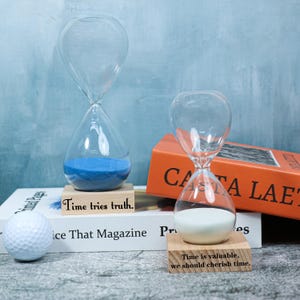 Könnte beinhalten: Zwei transparente Glas-Sanduhren mit blauem und weißem Sand, auf Holzblöcken mit Zitaten. Die Sanduhren stehen auf einem weißen Buch, eine Golfkugel ist sichtbar.
