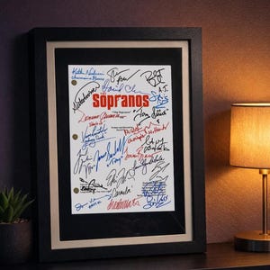 Könnte beinhalten: Gerahmter Druck einer Seite mit zahlreichen Unterschriften und dem Wort "Sopranos" in Rot. Die Unterschriften sind in verschiedenen Farben, darunter Blau und Rot. Der Rahmen ist schwarz mit einem goldenen Innenrand, präsentiert auf einer dunklen Oberfläche.