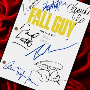 Pode incluir: Um roteiro branco com o título "THE FALL GUY" em amarelo e preto, com múltiplas assinaturas em tinta preta e azul. O roteiro está sobre um fundo de veludo vermelho. O texto "Written by Drew Pearce" e "Directed by David Leitch" também é visível.