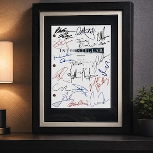 May include: A framed movie script page for Interstellar, featuring multiple signatures in black, blue, and red ink. The white paper has three punched holes on the left side and is encased in a black frame with a dark mat and a light inner border.