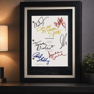 May include: Framed print featuring the title "Call Me By Your Name" in gold, with various signatures in red, blue, and black. The print is set against a cream background, framed in black, and mounted on a dark surface.