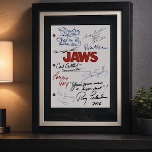 May include: Framed "Jaws" movie poster with signatures. The poster features the word "JAWS" in red, surrounded by handwritten signatures and quotes from the film. The frame is black with a white mount.