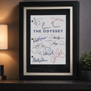 May include: A framed script cover titled THE ODYSSEY, featuring multiple handwritten signatures in black, blue, and red ink. The document has three circular punch holes on the left side and is encased in a black frame with a light-coloured matting.