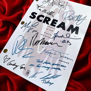 Könnte beinhalten: Ein weißes Drehbuch mit dem Titel "SCREAM" mit mehreren Unterschriften in blauer und schwarzer Tinte. Das Drehbuch ist mit goldenen Reißnägeln befestigt und liegt auf einer roten Samtoberfläche. Das Drehbuch enthält die Namen des Regisseurs Wes Craven und des Autors Kevin Williamson.