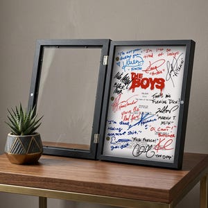 Puede incluir: Un marco de exhibici&oacute;n negro con un fondo blanco, con firmas y el texto rojo "THE BOYS". El marco est&aacute; abierto, revelando las firmas en tinta azul y roja. A la izquierda hay una peque&ntilde;a suculenta en una maceta geom&eacute;trica.
