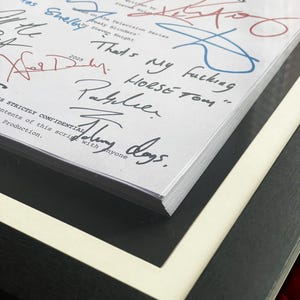 Puede incluir: Un guion enmarcado de "Peaky Blinders: The Immortal Man" con m&uacute;ltiples firmas en tinta azul y roja. El t&iacute;tulo y el nombre del autor son visibles. El marco es negro con un borde blanco, y el guion est&aacute; asegurado con un cierre dorado.