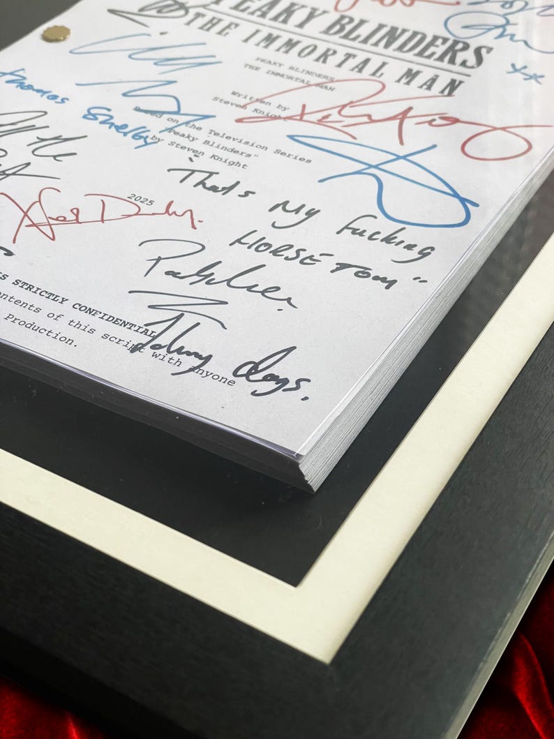 Puede incluir: Un guion enmarcado de "Peaky Blinders: The Immortal Man" con m&uacute;ltiples firmas en tinta azul y roja. El t&iacute;tulo y el nombre del autor son visibles. El marco es negro con un borde blanco, y el guion est&aacute; asegurado con un cierre dorado.