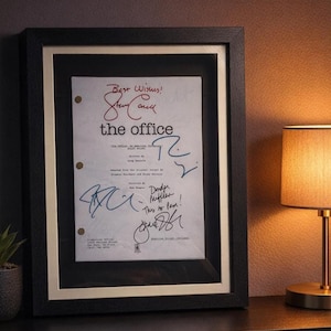 Puede incluir: Guion enmarcado de "The Office" con firmas. El guion está en un marco negro con un paspartú blanco. El guion tiene el título "the office" y las palabras "Best Wishes!" en tinta roja. Se ven varias firmas.