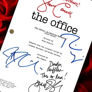 Puede incluir: Guion de rodaje blanco de "The Office" con múltiples firmas en tinta azul y negra. El título "the office" está en negro, con "Best Wishes! Steve Carell" en rojo. Fondo de tela de terciopelo rojo.