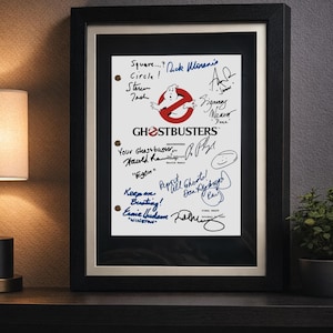 May include: Framed Ghostbusters movie script draft with signatures. The framed artwork features the Ghostbusters logo and text, with handwritten notes and signatures in blue ink. The frame is black.