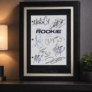 May include: A framed script cover for The Rookie, featuring the title in dark blue text and multiple black and blue ink signatures. The document is set against a white background with two circular punch holes on the left side, encased in a black frame.