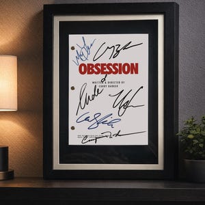 May include: A framed script cover titled OBSESSION, written and directed by Curry Barker, featuring multiple handwritten signatures in black and blue ink. The document is displayed in a black frame with a white mat and has three visible binder holes.