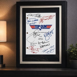 Op de afbeelding: Ingelijst Top Gun-filmscript met meerdere handtekeningen. Het script bevat het Top Gun-logo in rood, wit en blauw. De lijst is zwart en de passe-partout is gebroken wit. Het script wordt weergegeven op een donkere ondergrond.
