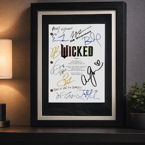 May include: A framed script cover for the musical Wicked, featuring the title in stylised green and black lettering. The white page includes multiple handwritten signatures in blue, black, and gold ink, along with the text Screenplay by Winnie Holzman and Dana Fox.