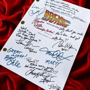 Puede incluir: Una página de guion blanca con el logo de "Regreso al Futuro" y firmas manuscritas en varios colores. El guion está sobre un fondo de terciopelo rojo. El texto incluye "Marty" y "Great Scott!"