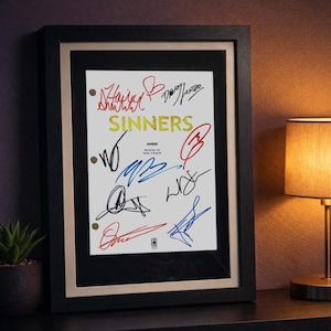 May include: Framed artwork featuring the word "SINNERS" in yellow, with multiple colorful signatures. The artwork is in a black frame with a cream-colored mat, displayed on a dark surface with a lamp and plant in the background.