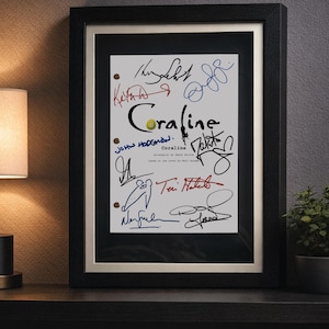 May include: Framed artwork featuring the title "Coraline" with multiple signatures in various colors. The signatures are on a white background, framed in black with a gold inner border. The artwork is displayed on a dark shelf.