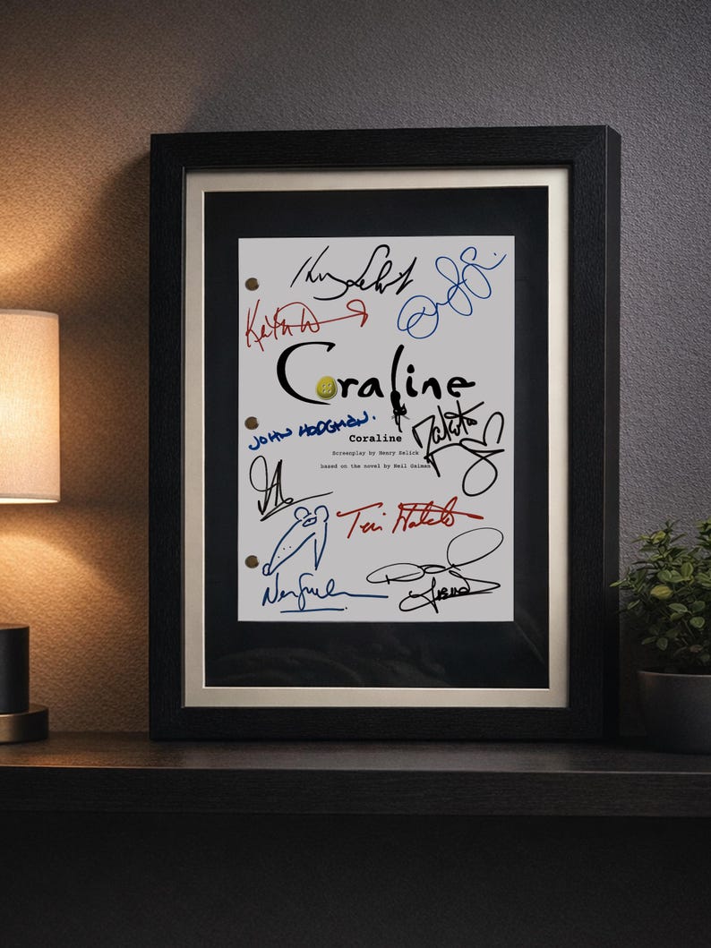 May include: Framed artwork featuring the title "Coraline" with multiple signatures in various colors. The signatures are on a white background, framed in black with a gold inner border. The artwork is displayed on a dark shelf.