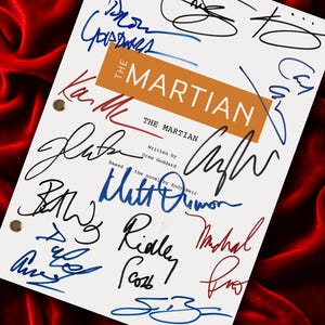 May include: A white screenplay cover for The Martian, written by Drew Goddard and based on the novel by Andy Weir, featuring a central orange rectangular block with the title and numerous handwritten signatures in black, blue, and red ink.